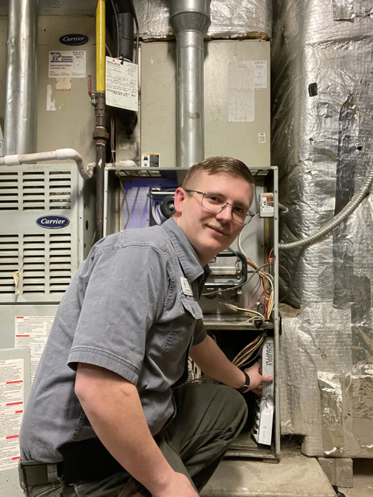 A man in a gray shirt kneeling beside a furnace, working diligently.