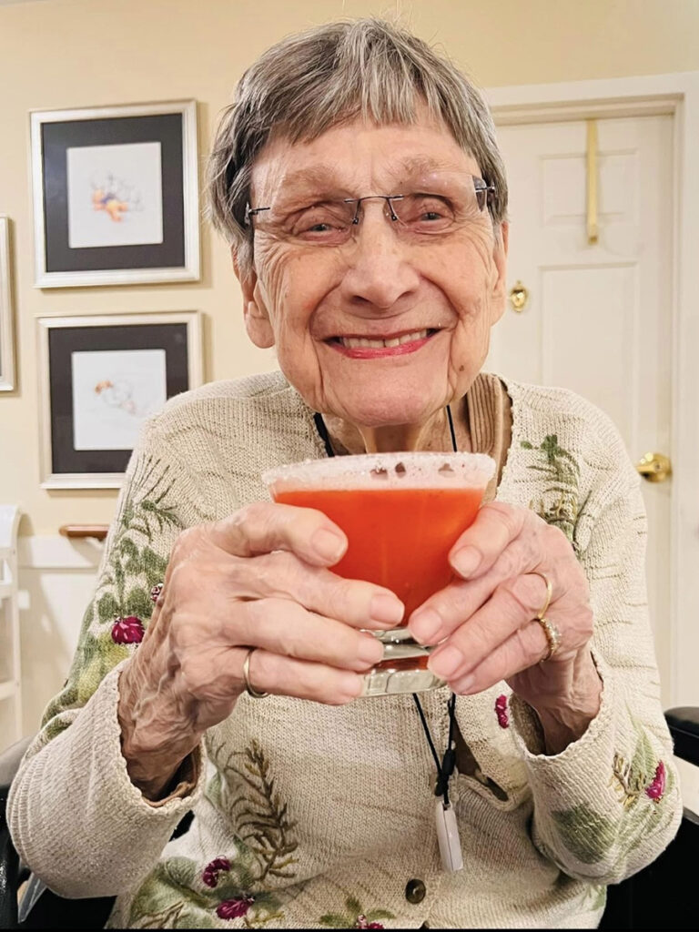 This is a joyful current photo of Joan smiling in her beige button-up sweater with embroidered flowers and green leaves. She is holding a cup with a fun reddish orange drink.