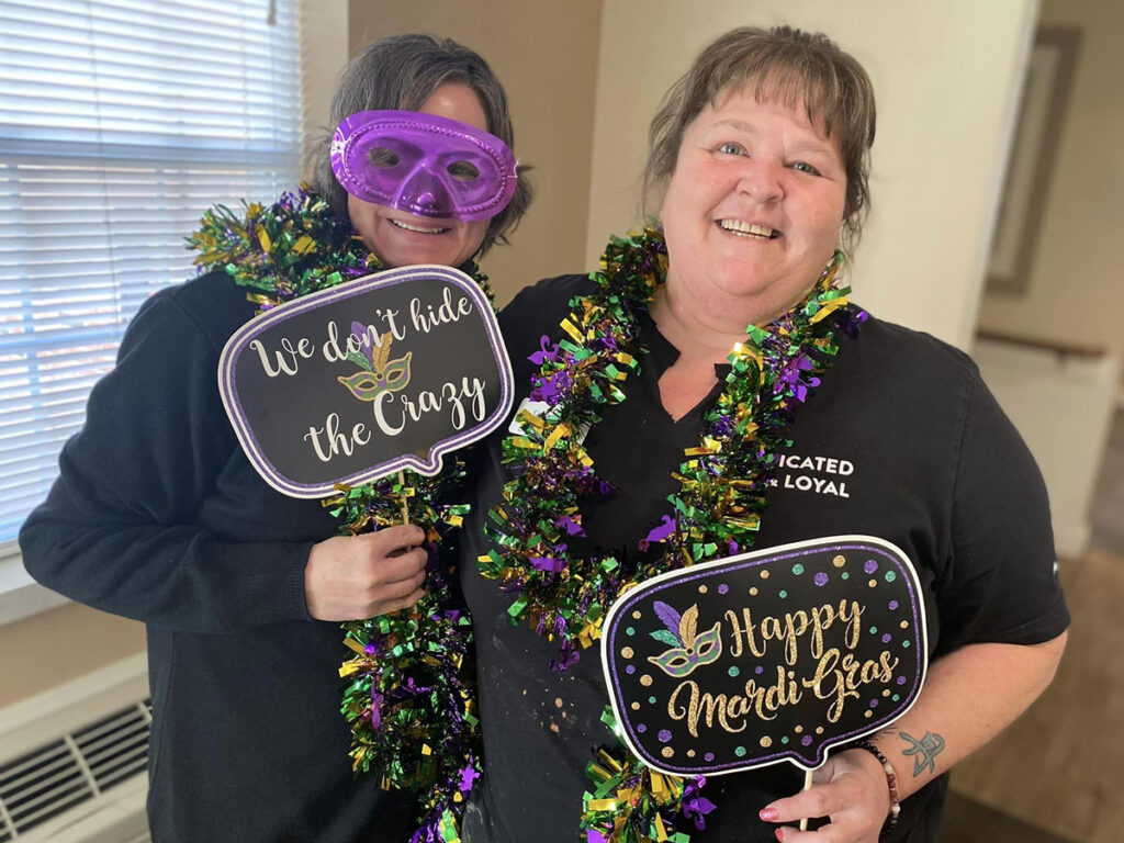 Two senior living employees in black shirts and masks holding up signs at a Mardi Gras event.