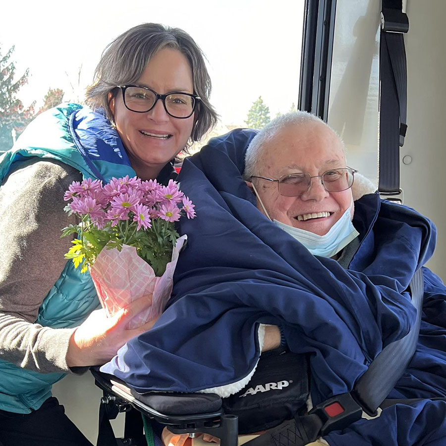 Experience Mom & Pop Small Business Day with Wendy, our Resident Services Director, at The Olympic Diner. She is holding bright pink flowers as an elderly man in a wheelchair is greeted at the diner.