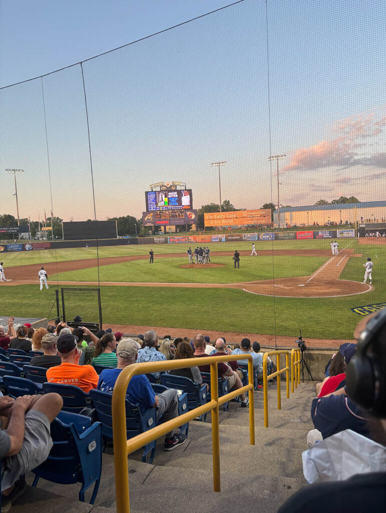 View of a baseball game from the stands, with players on the field and a large scoreboard in the background during sunset.