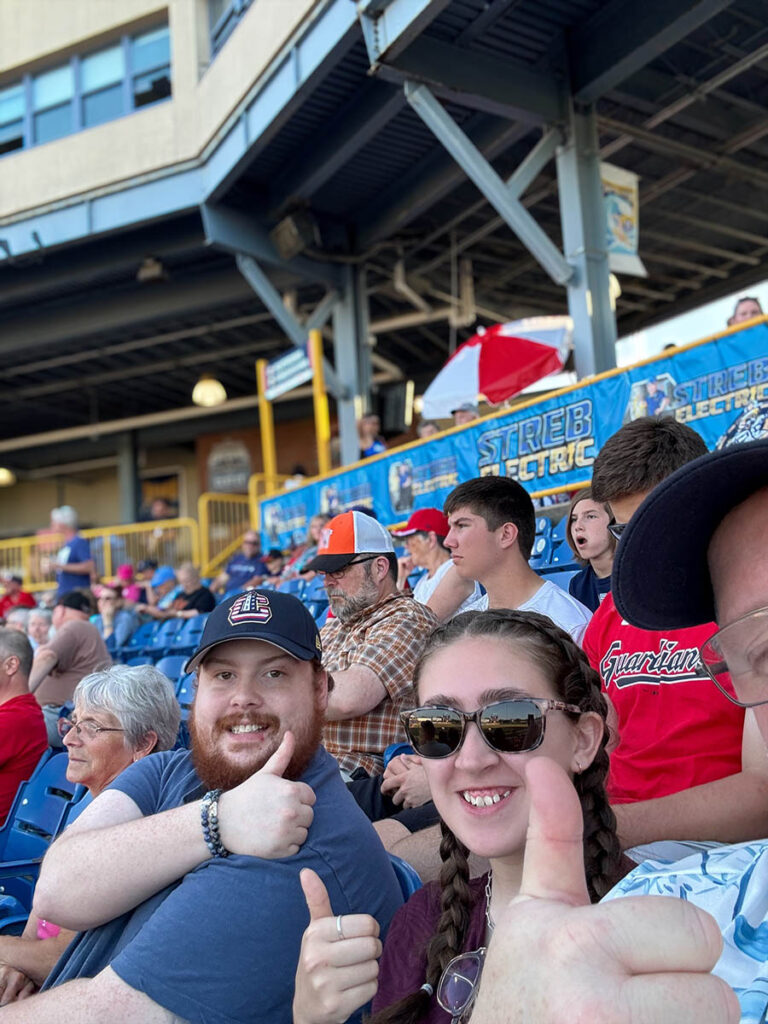 Fans at a baseball game giving thumbs-up, sitting in stadium seats with other spectators in the background.