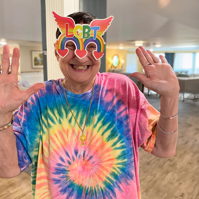 Senior lady in a vibrant tie-dye shirt joyfully poses with hands up, wearing colorful LGBT glasses with heart decorations in a cheerful room.