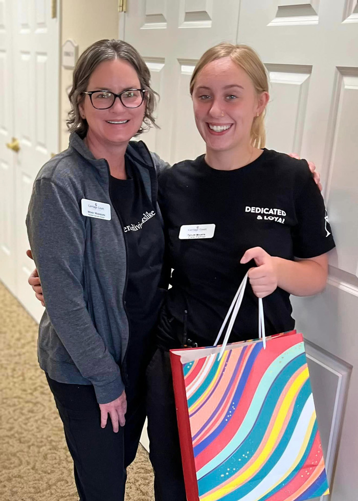 Two staff members at a senior living community smile together as they celebrate one of their Care Partners going back to school. One holds a colorful gift bag, creating a supportive and encouraging atmosphere.