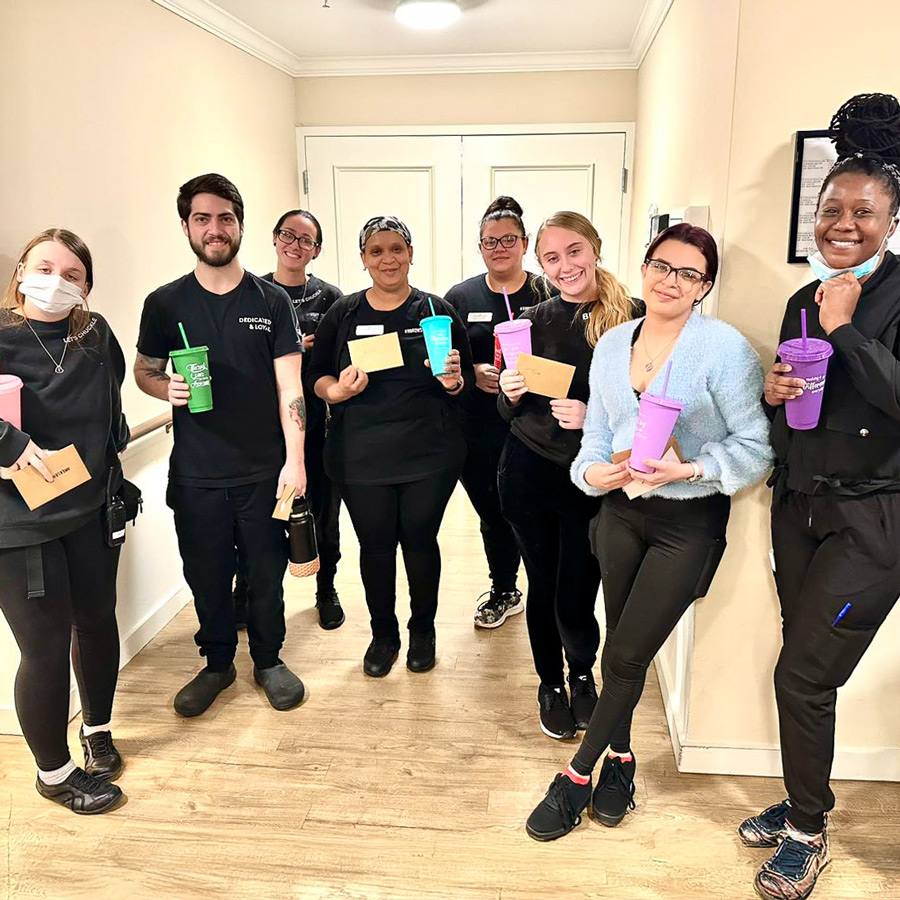 A group of staff members, likely nurses, at a senior living community stand together, smiling, and holding colorful tumblers and envelopes, celebrating Nurses Week in a warm and appreciative atmosphere.