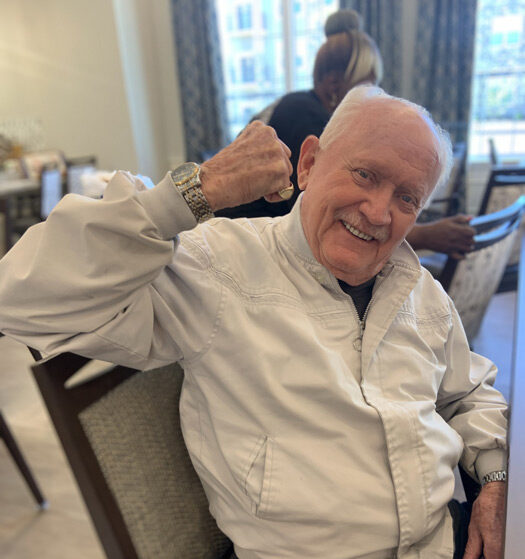 Smiling elderly man wearing a white jacket, sitting on a chair and playfully flexing his arm in a dining area.