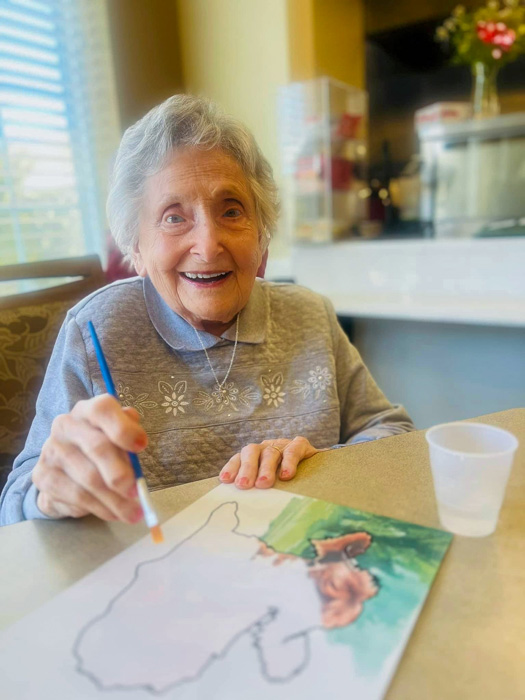 Elderly woman smiling and painting a picture at a table at Cedar Trails Senior Living.