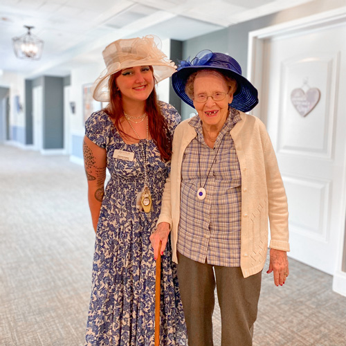 In a hallway at a senior living community, a senior woman wearing a blue hat and plaid shirt stands with a young woman in a floral dress and white hat, both smiling and about to head to a special event.