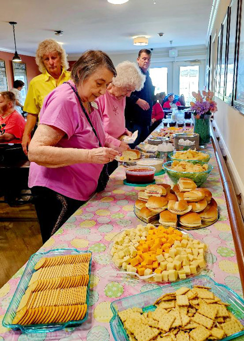 Senior residents enjoy a vibrant buffet filled with sandwiches, cheese, crackers, and snacks during a cheerful community event with friends and neighbors.