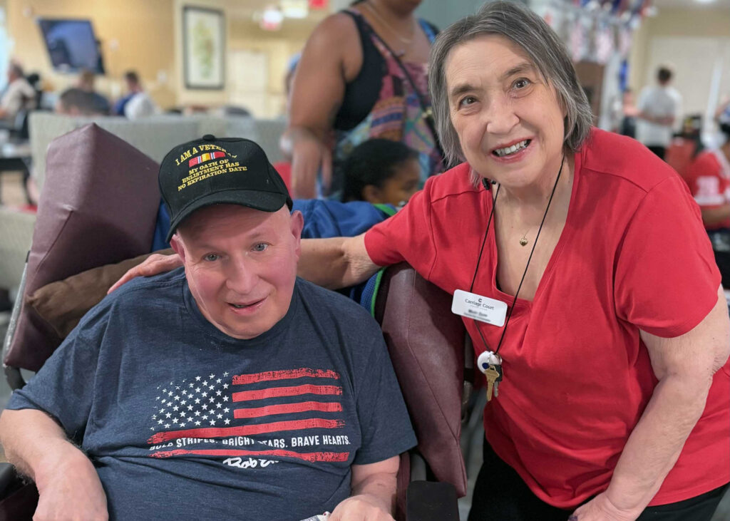Senior man in a veteran hat and patriotic shirt sits beside a smiling woman in a red top at Carriage Court Senior Living, celebrating a community event together.