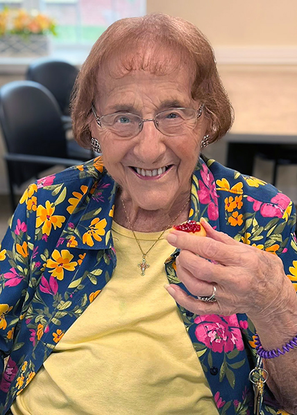 A senior woman in a brightly colored outfit smiles, holding up a jam-covered snack at Carriage Court.