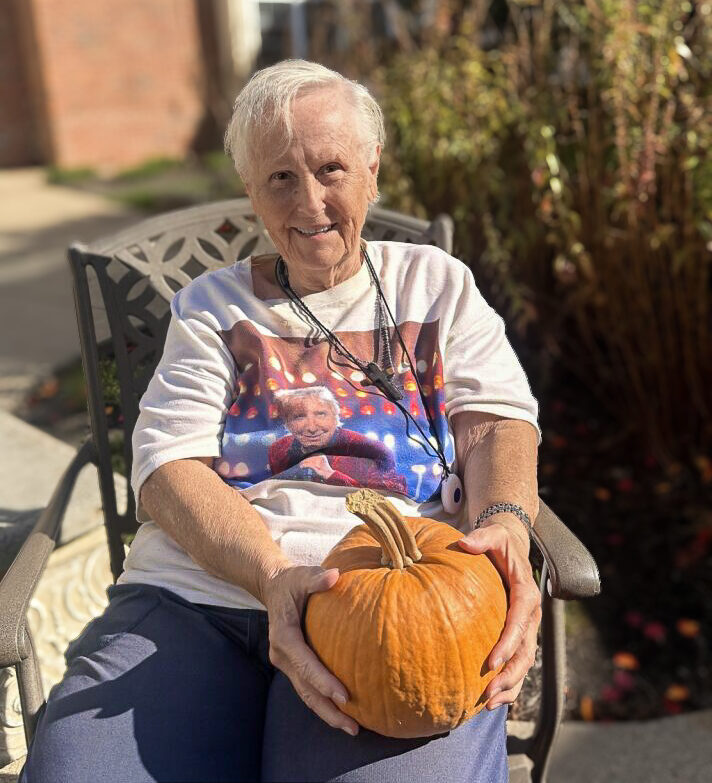 A resident at Carriage Court Senior Living smiles, holding a pumpkin, during a sunny Autumn day.