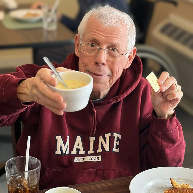 A smiling senior man raises a bowl of chef-prepared soup.