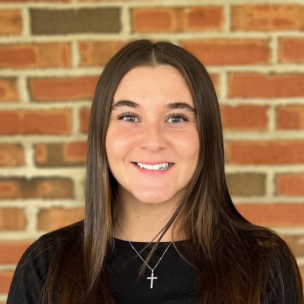 Raven Steele, Senior Living Director at Carriage Court Senior Living, smiling in a professional headshot, wearing a black top and cross necklace, standing in front of a brick wall background.
