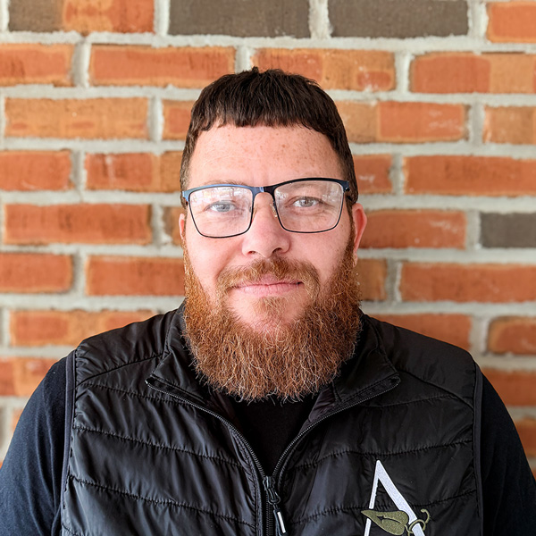 Steve Jordan, Plant Operations Director at Carriage Court Senior Living, looking at the camera in a professional headshot with short dark hair, glasses, a full red beard, a black vest, and a red brick wall background.