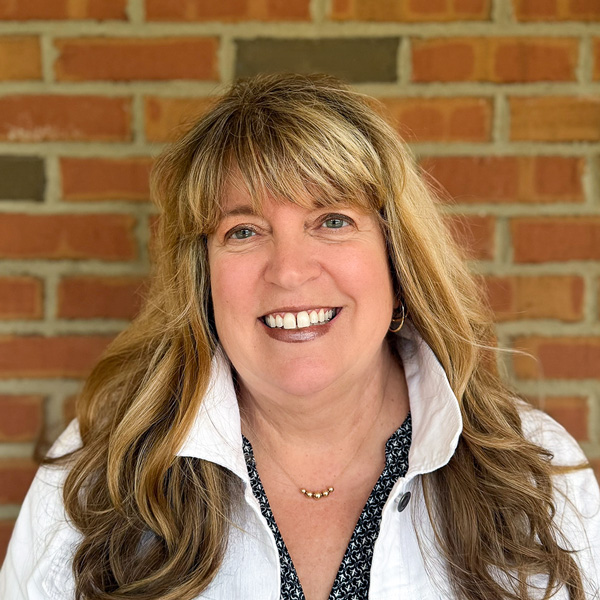 Lisa Hord, Executive Director at Carriage Court Senior Living, smiling in a professional headshot with long blonde hair and bangs, a white jacket, and a red brick wall background.