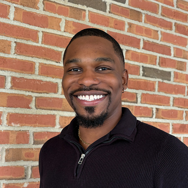 Michael “Mike” Kusi, Business Operations Director at Carriage Court Senior Living, smiling in a professional headshot with closely cropped hair, a trimmed goatee, a dark quarter-zip sweater, and a red brick wall background.